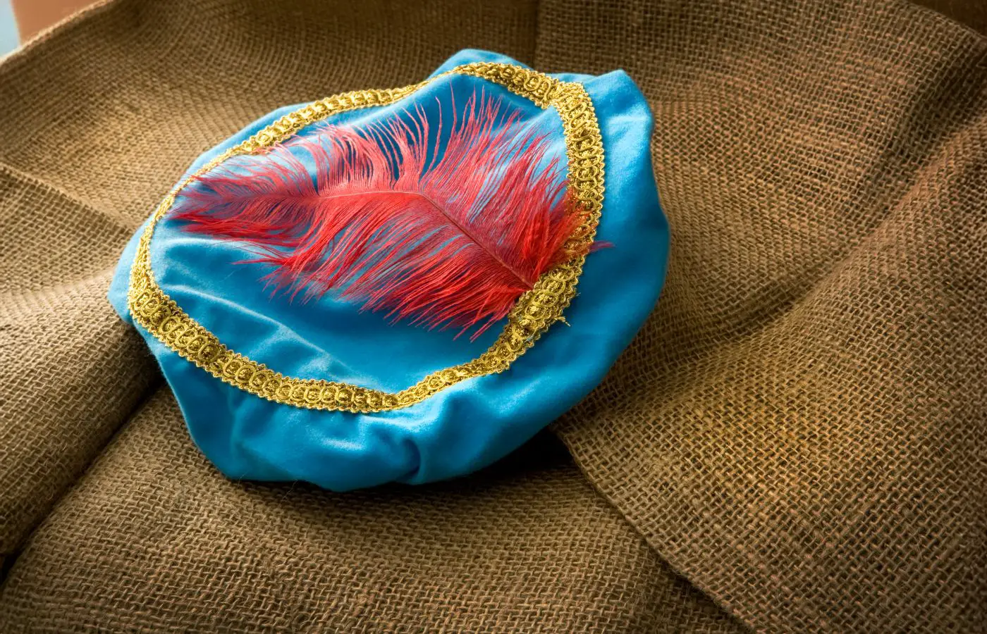 Traditionele hoed van Zwarte Piet in blauwe satijn, versierd met een rode veer en gouden rand, liggend op een juten zak, symbool van het Sinterklaasfeest.