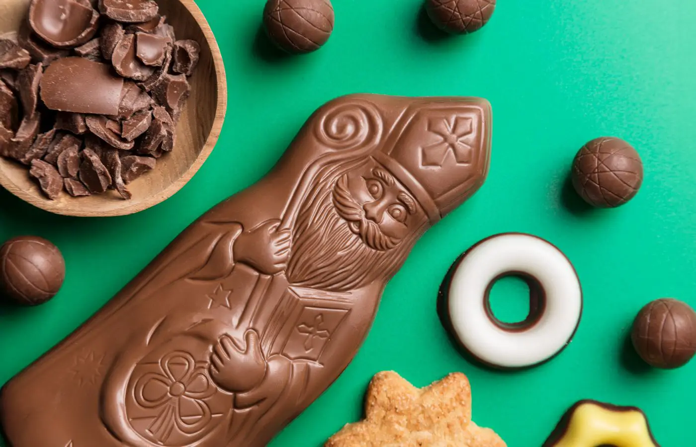 Chocoladefiguur van Sinterklaas met feestelijke decoratie, omringd door chocoladeschaafsel, ronde snoepjes en koekjes op een groene achtergrond.