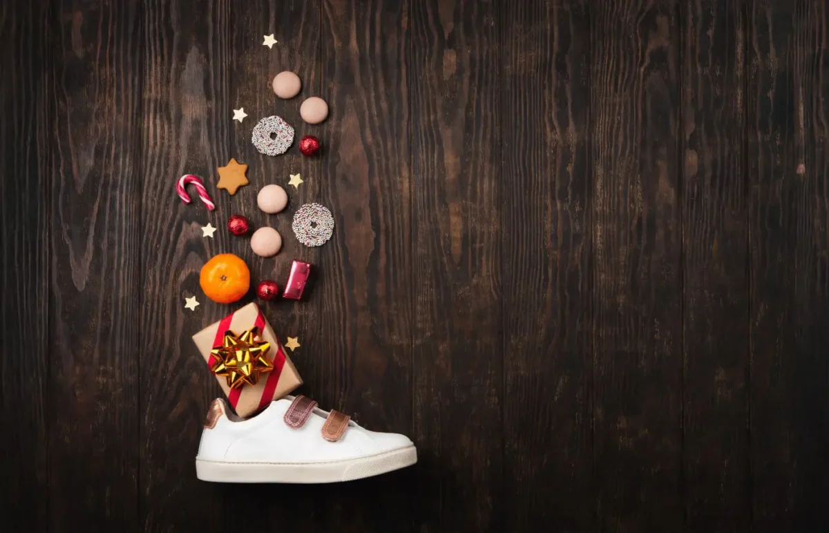 Witte schoen gevuld met Sinterklaasgeschenken zoals snoep, mandarijnen, cadeautjes en decoratieve sterren, op een donkere houten achtergrond.